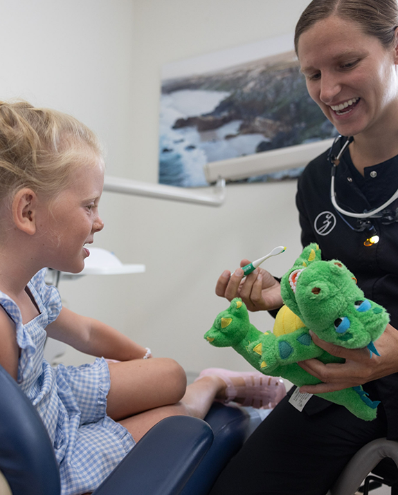 Young girl at a dental visit watching a dental team member brush the teeth of a stuffed crocodile